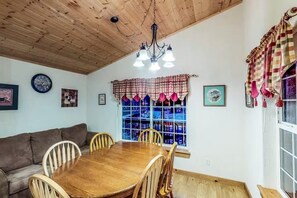 Ein Essbereich mit Holztisch und Stühlen, einer Kristallleuchter, einem Fenster mit Vorhängen sowie einem Sofa.