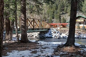 A wooden bridge over a river with snow on the ground, surrounded by trees and a cabin with a red roof. - Gressoney Family Retreat | Pond & Hot Tub (South Fork)