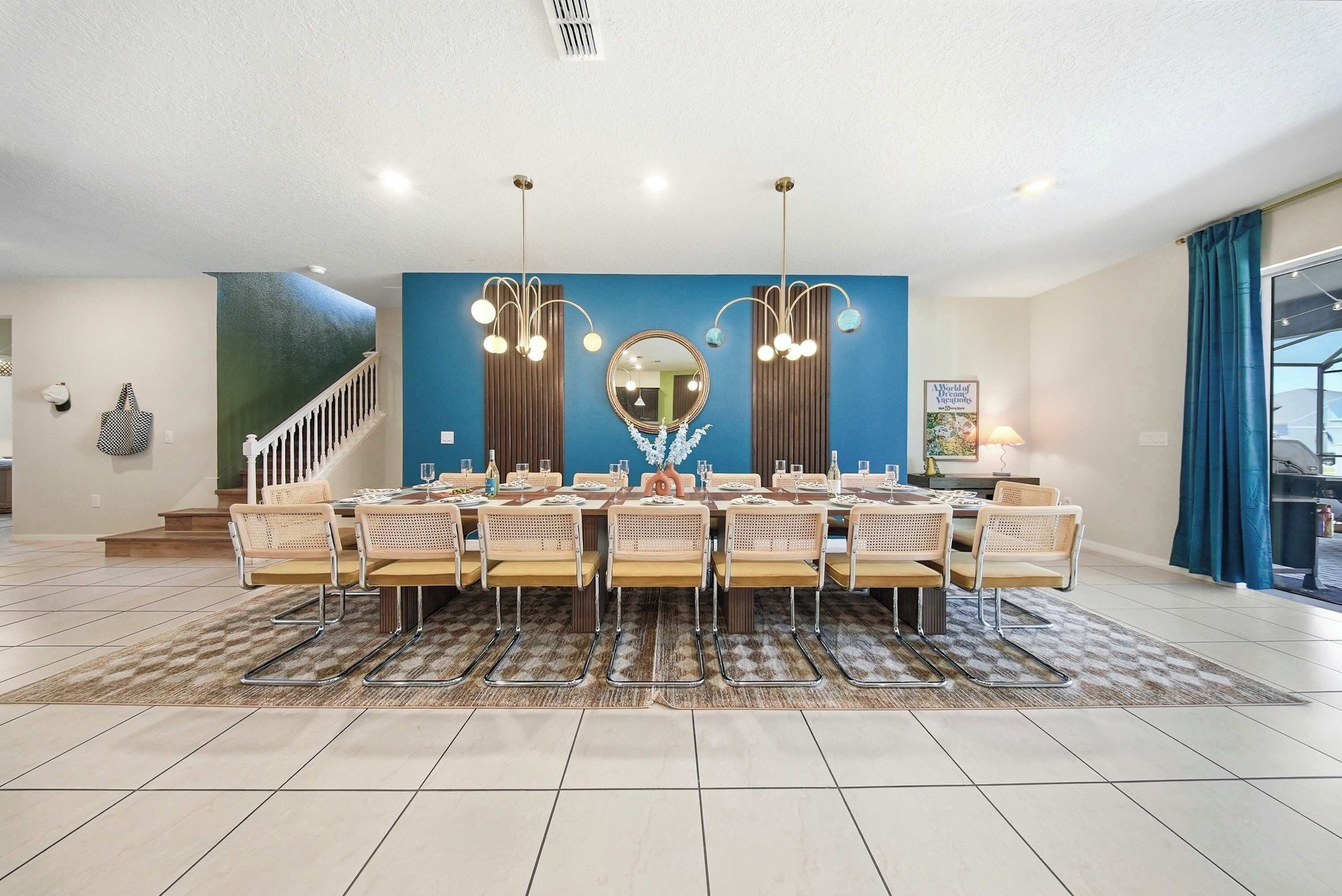Un espace repas avec une longue table dressée pour six personnes, flanqué de deux lustres fixés au mur, d’un grand miroir et d’un escalier menant à un niveau supérieur.