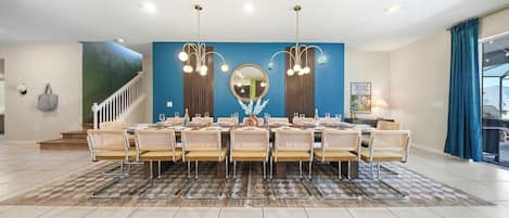 A dining area with a long table set for six, flanked by two wall-mounted chandeliers, a large mirror, and a staircase leading to an upper level.