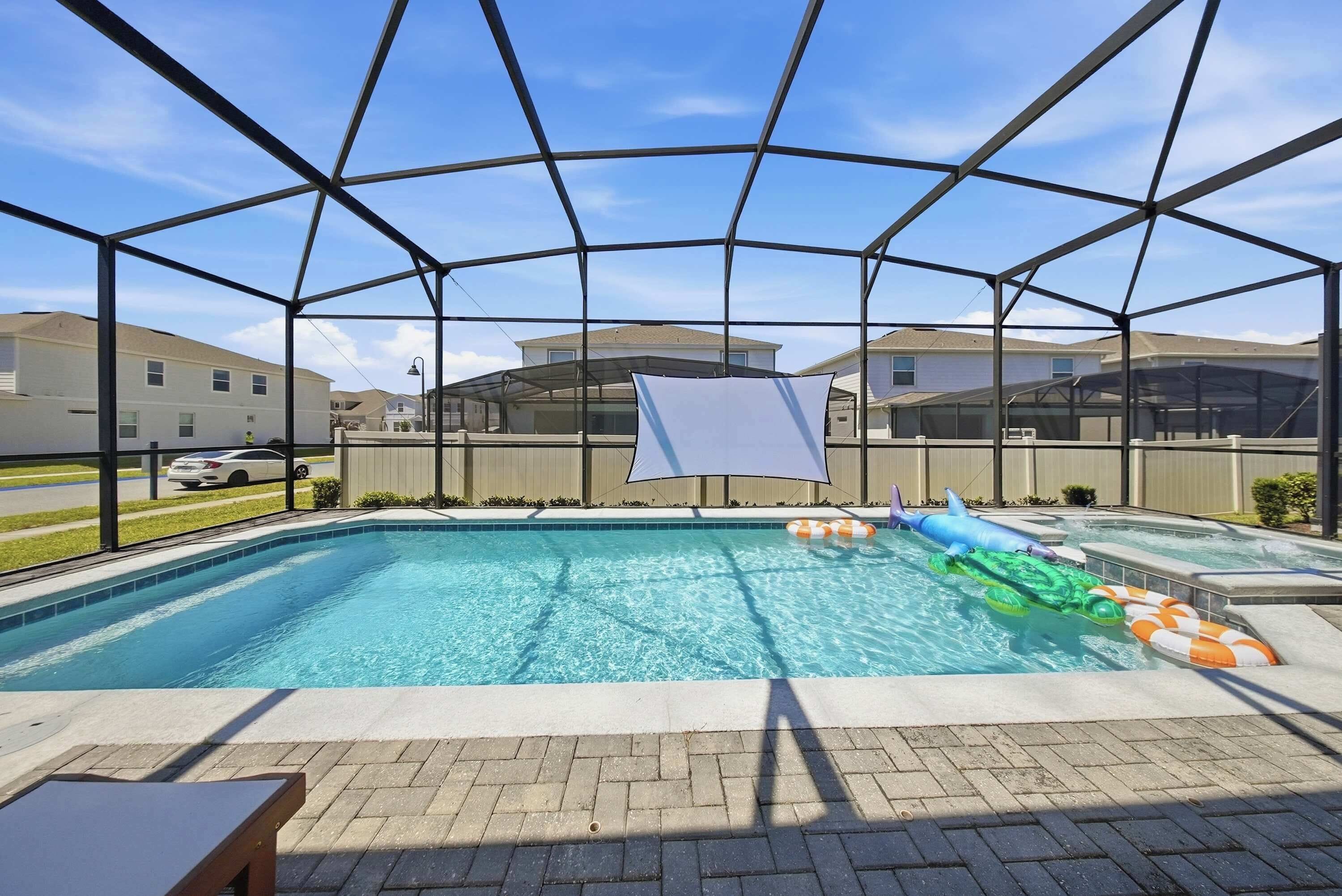 A swimming pool with a safety net enclosure, surrounded by a paved deck and a view of residential buildings.