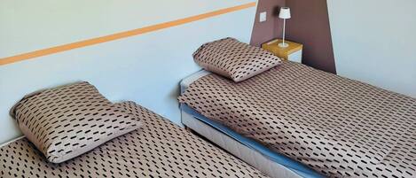 A hotel room with two single beds, a wall-mounted fan, and a nightstand with a lamp.