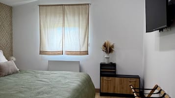 Une chambre à coucher avec un lit, un téléviseur fixé au mur, une fenêtre avec des rideaux, une table de chevet avec un vase et un plancher en bois.