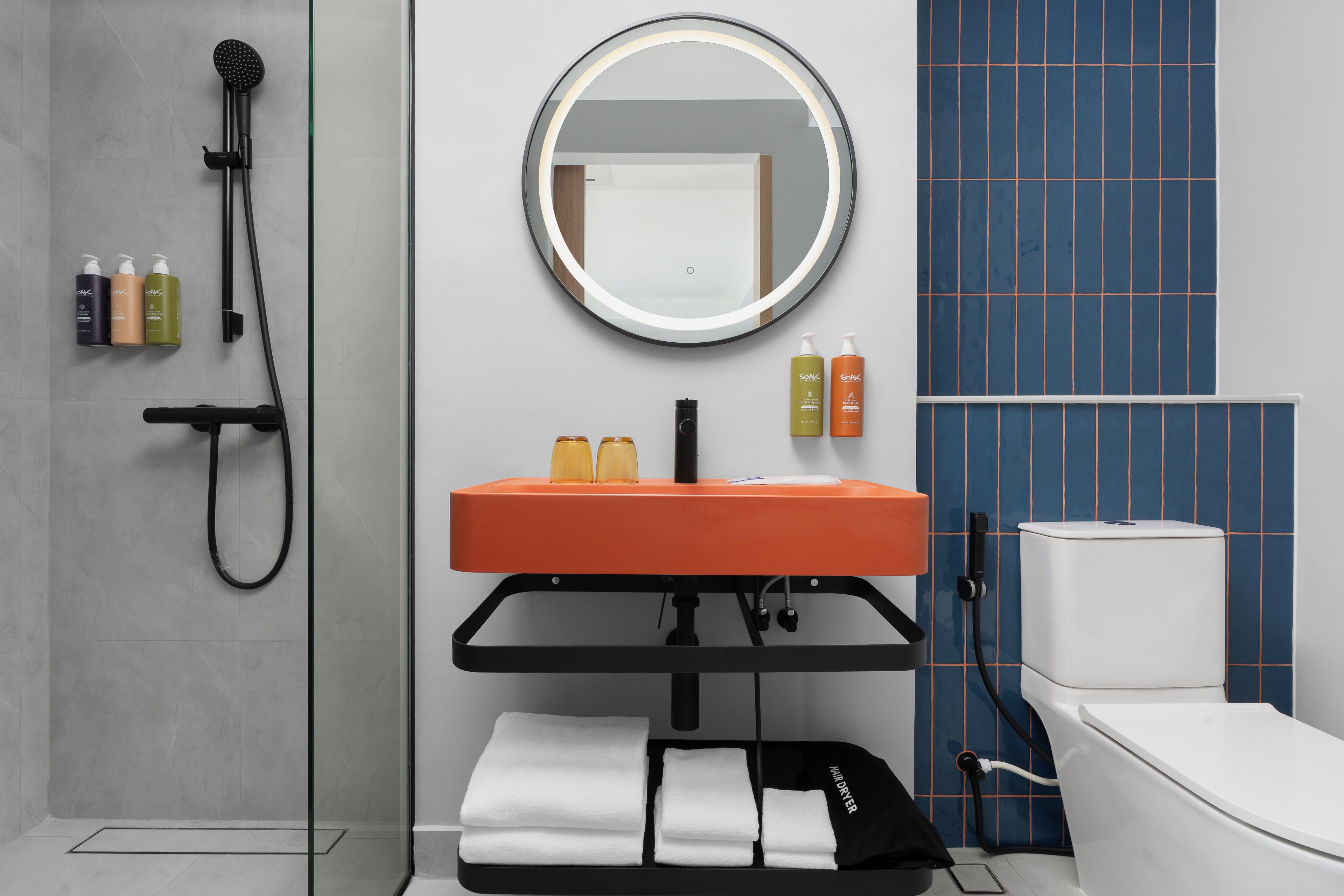 Une salle de bain moderne avec une douche à parois de verre, un grand miroir rond avec éclairage intégré, un lavabo noir et des toilettes.