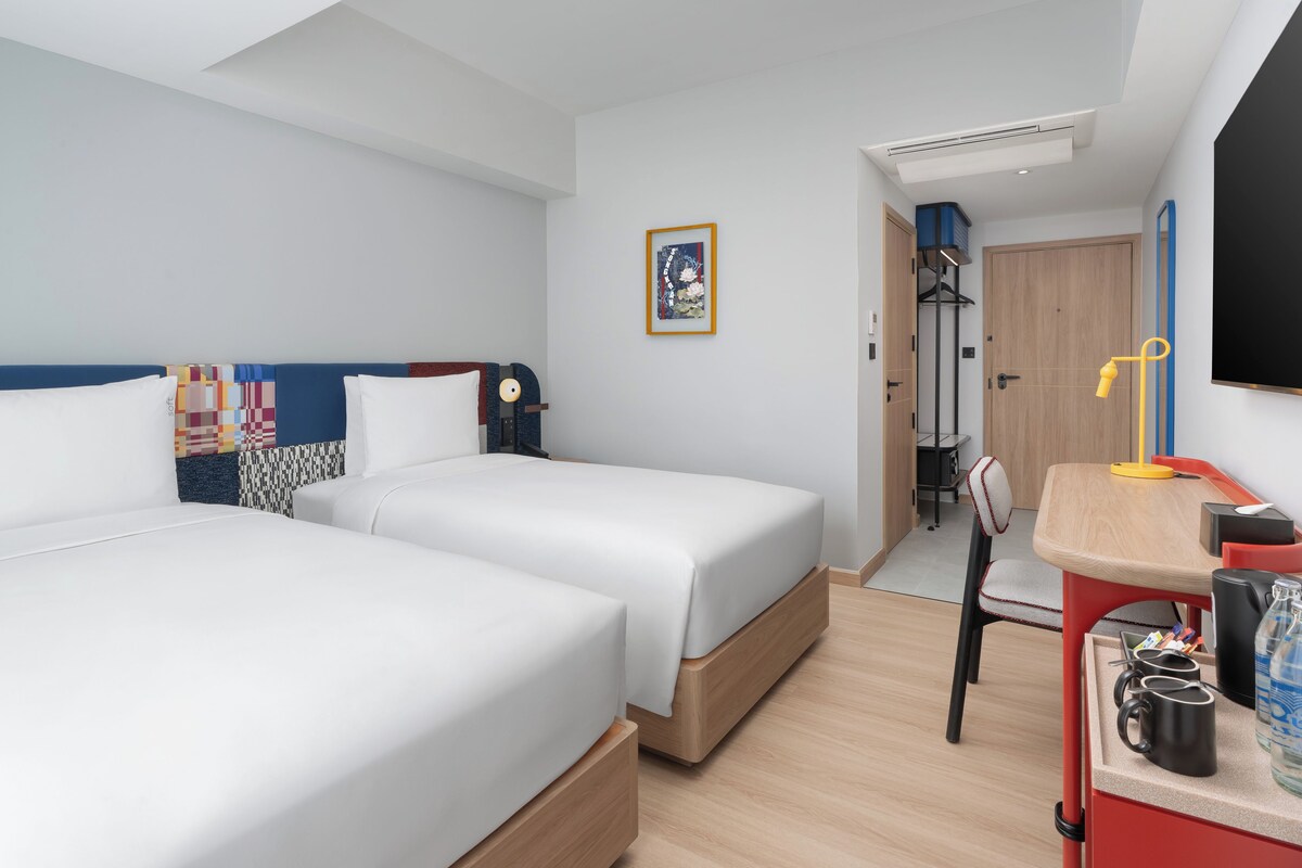 A hotel room with two beds, a desk with a chair, a TV, and a door leading to a bathroom.