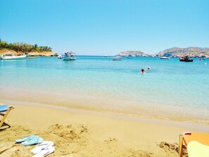 Ein Strand mit klarem türkisfarbenem Wasser, Booten und einem sandigen Ufer mit Flip-Flops und einer Liege.
