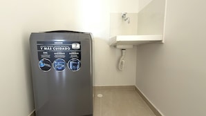 Una lavadora compacta gris, fijada a la pared, con un panel de control que incluye pantalla táctil y tres indicadores circulares.