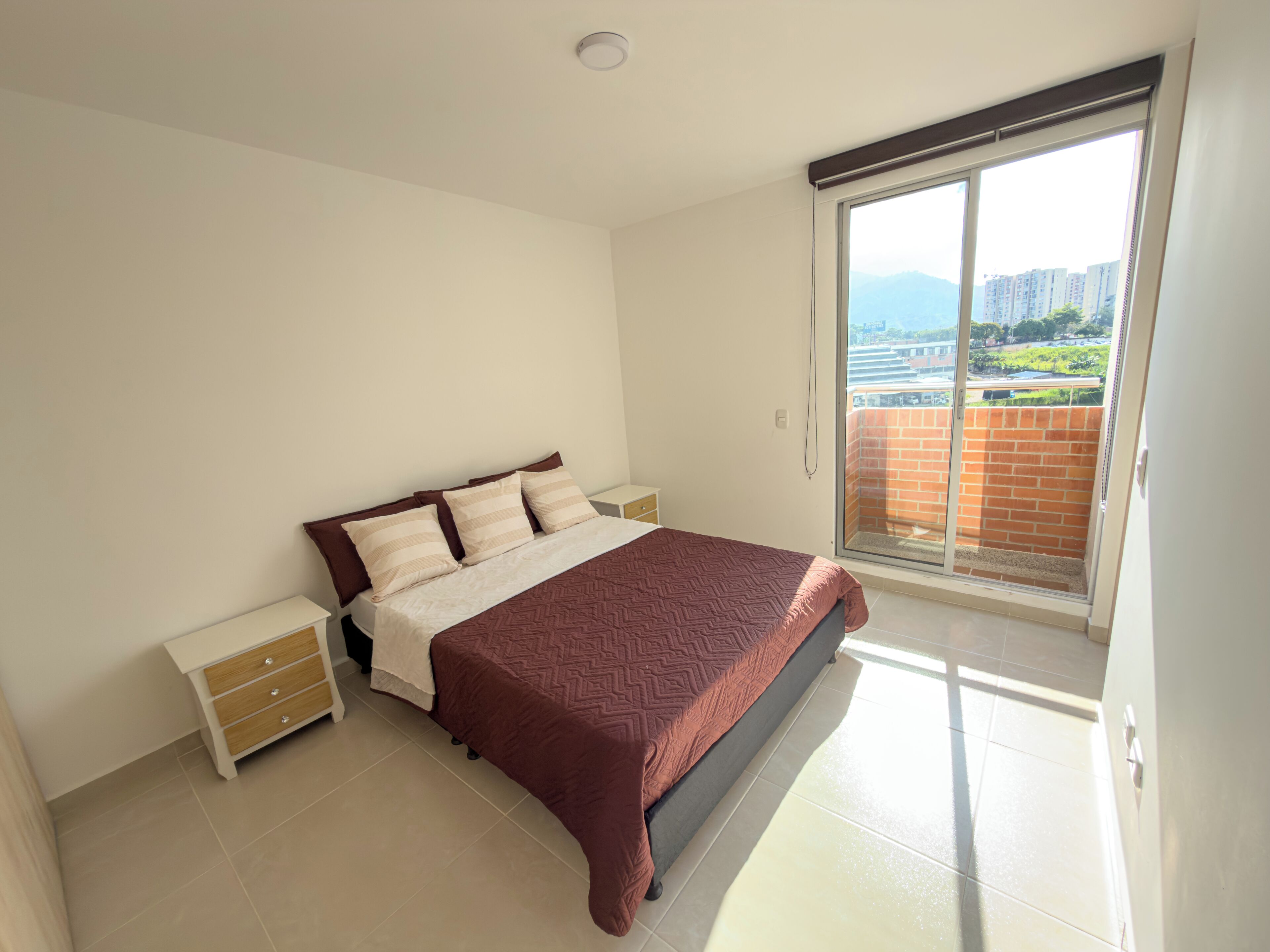 A bedroom with a bed, nightstands, a sliding glass door to a balcony, and a view of buildings and greenery.
