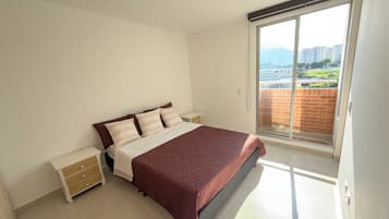 A bedroom with a bed, nightstands, a sliding glass door to a balcony, and a view of buildings and greenery.