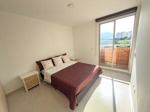 Un dormitorio con cama, mesitas de noche, puerta corrediza a un balcón y vistas a edificios y vegetación.