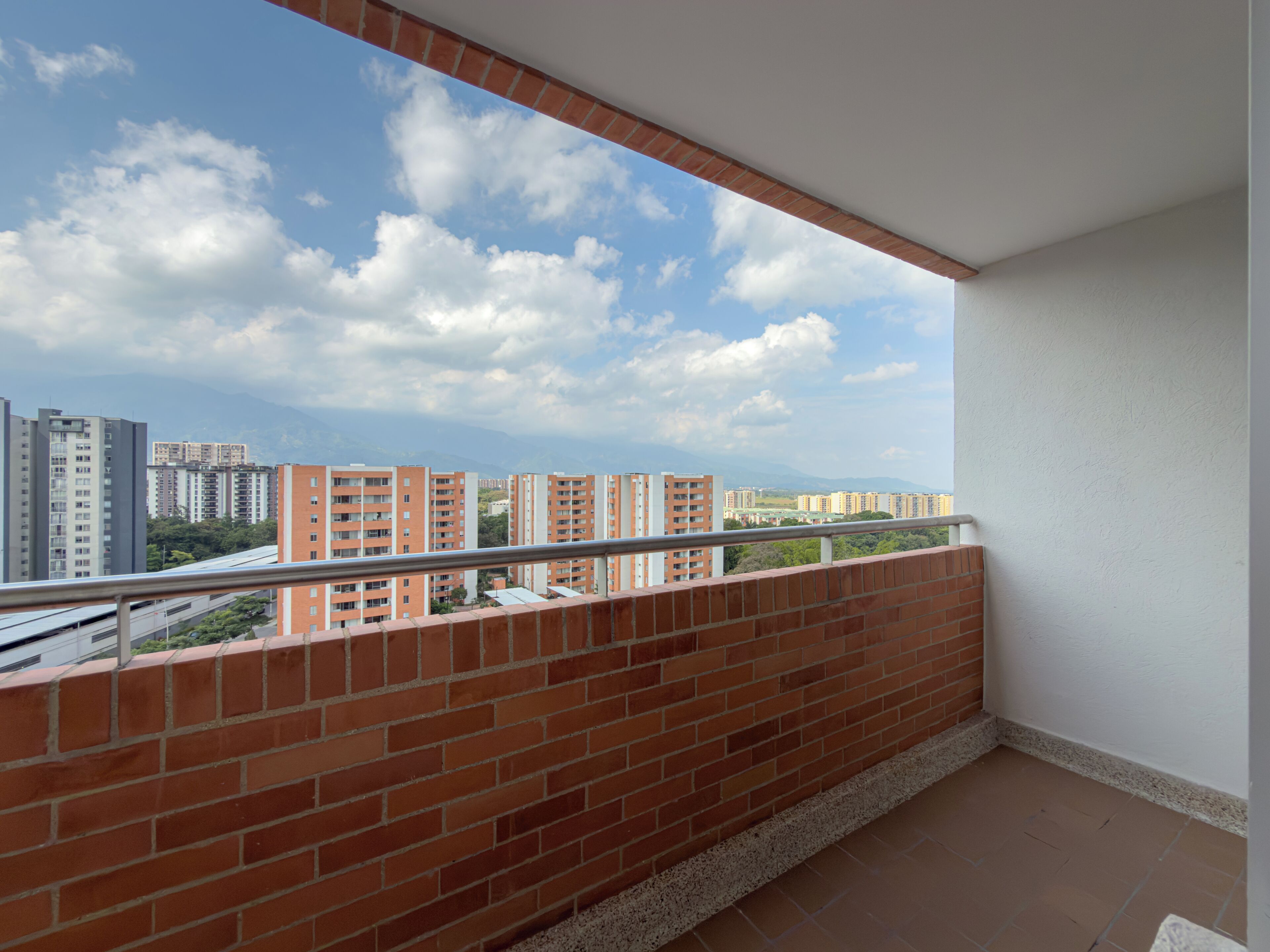 A balcony with a brick wall and metal railing, overlooking a cityscape with multiple high-rise buildings and a clear sky.