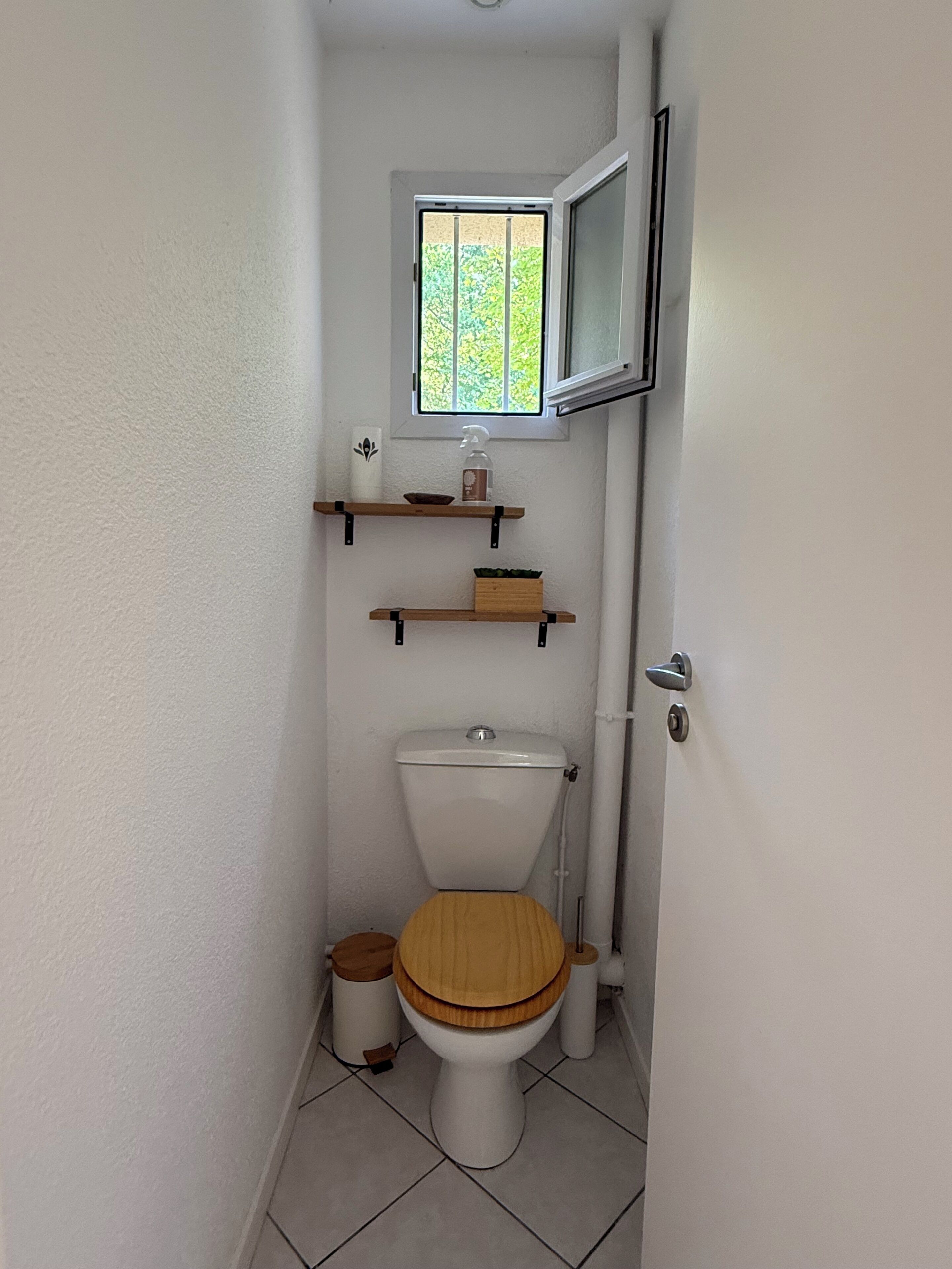 Baño pequeño y funcional con inodoro, estantes de madera, una ventana y un televisor sobre el inodoro.