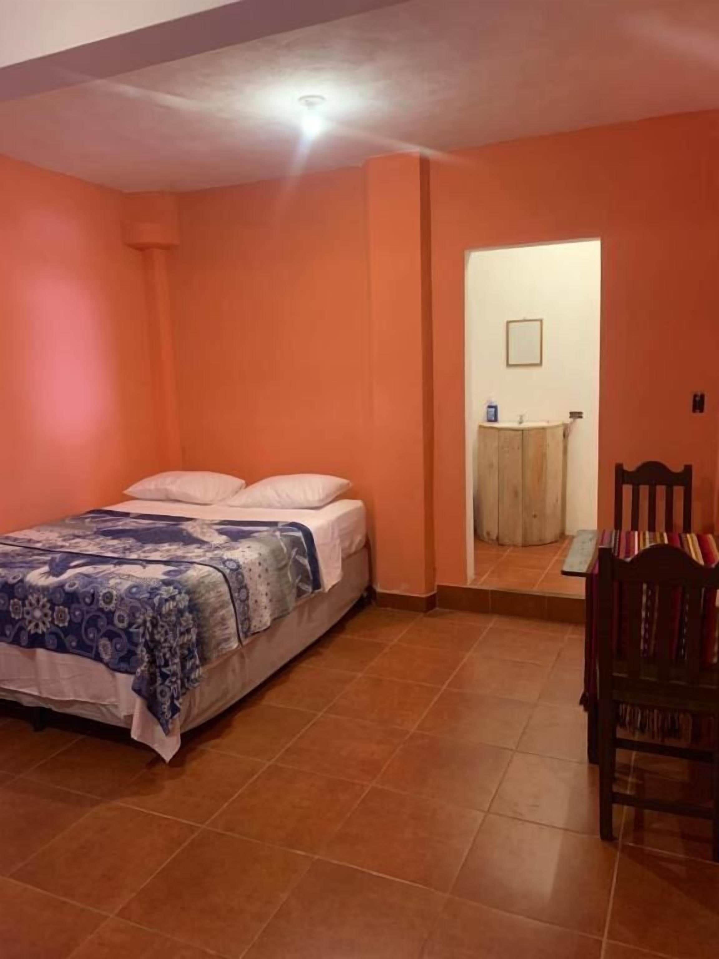 A bedroom with a bed, a wooden chair, a small table, and a bathroom visible through an open door.