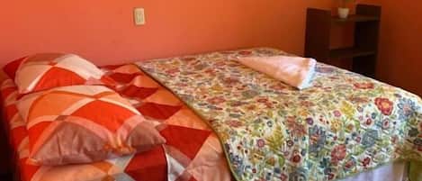 Una cama con una colcha estampada y almohadas, una mesita de noche con una planta y una alfombra en el suelo.
