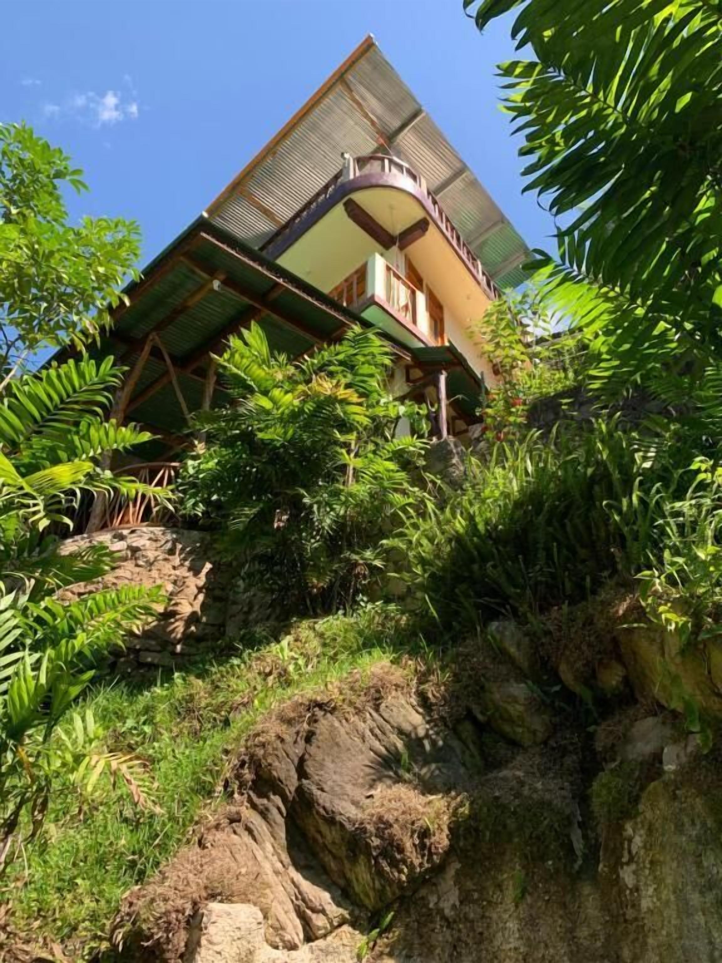 A building with a sloped roof, surrounded by lush greenery and a rocky landscape.