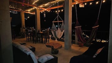 Un área de estar al aire libre con hamacas, mesas y sillas, bajo una estructura cubierta con luces colgantes.