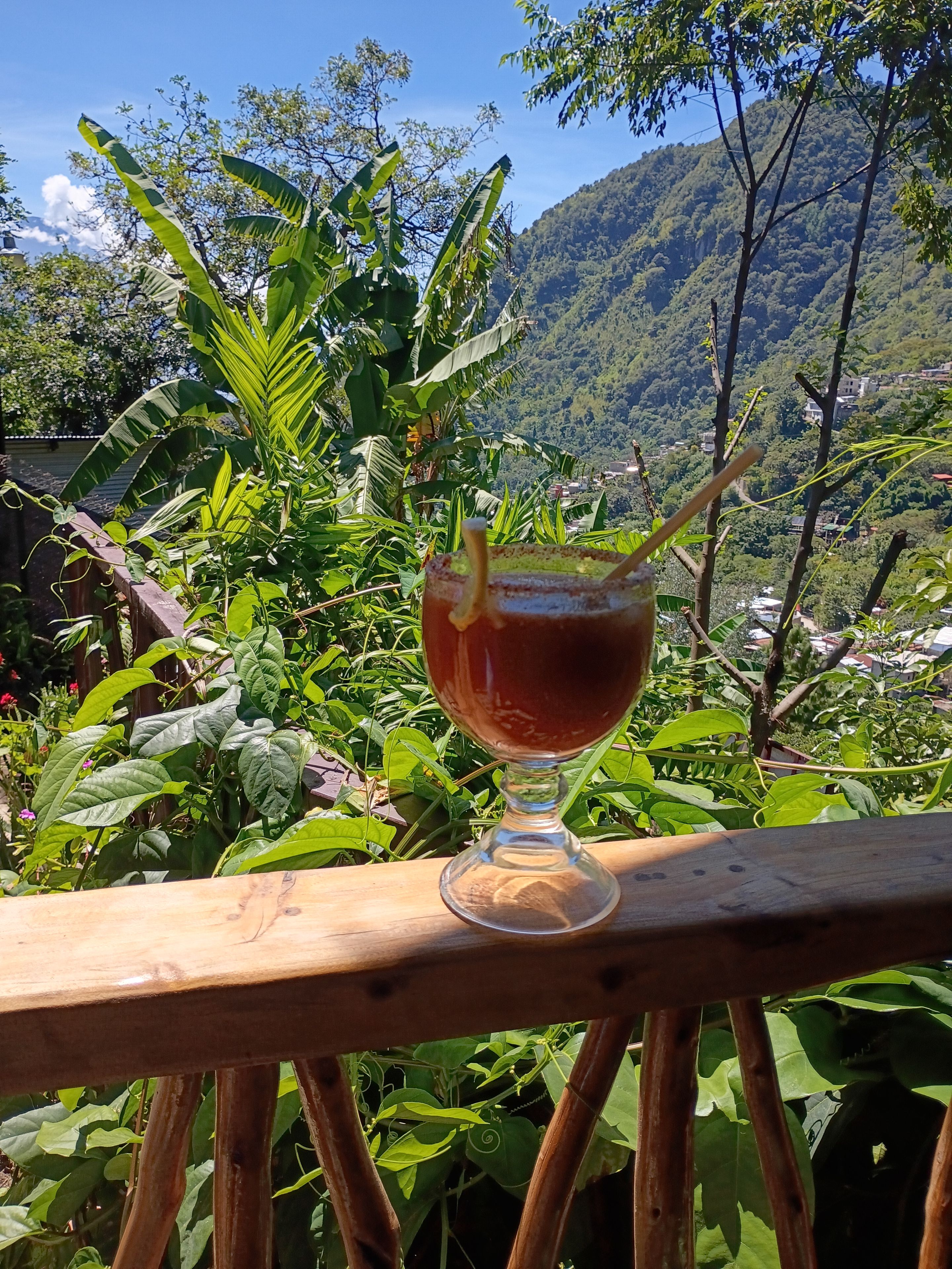 A glass of beverage on a wooden railing with a mountainous landscape in the background.