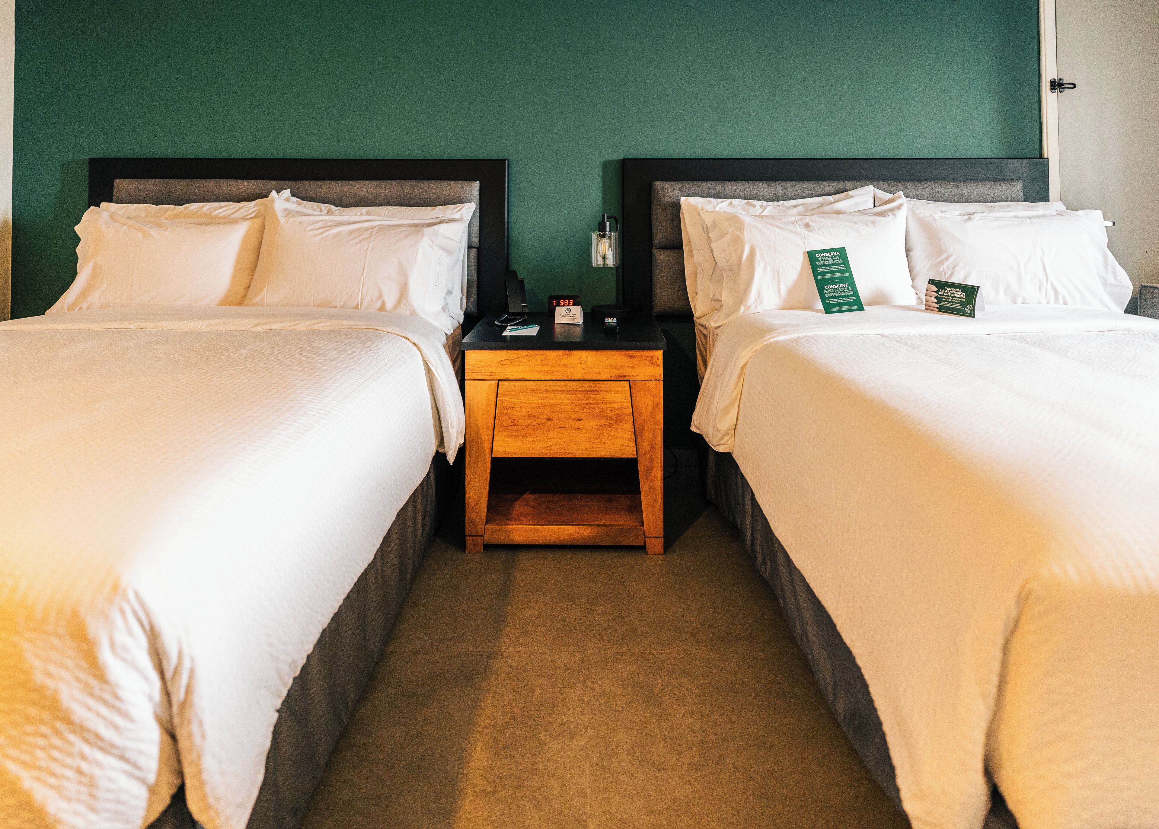 Quarto de hotel com duas camas, uma mesinha de cabeceira de madeira e uma parede verde.