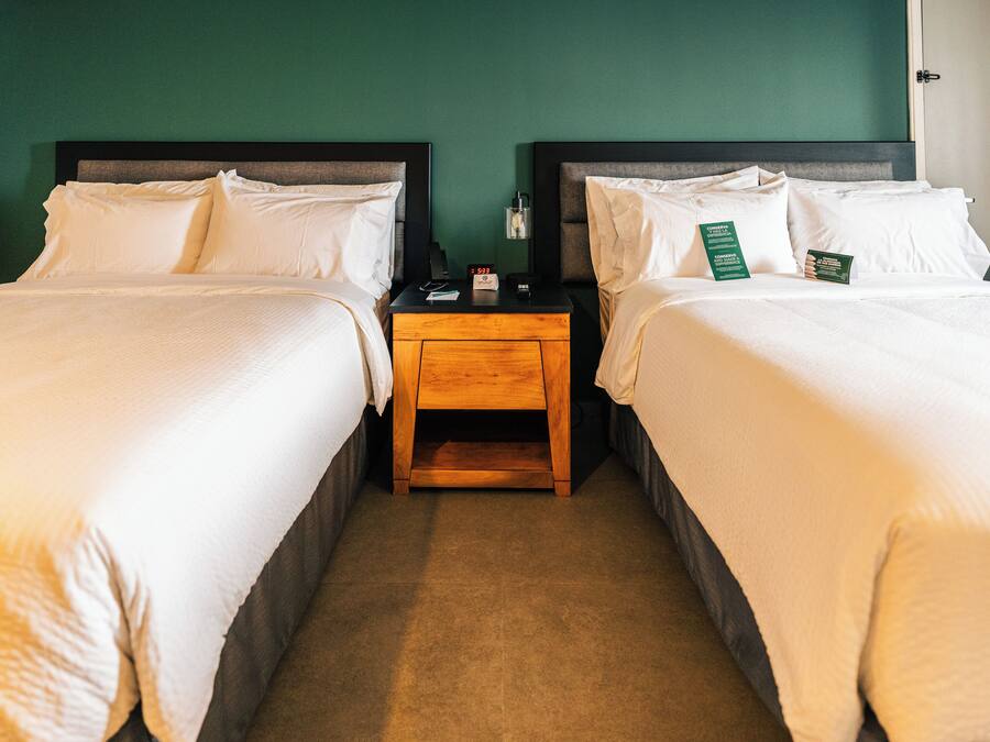 Quarto de hotel com duas camas, uma mesinha de cabeceira de madeira e uma parede verde.