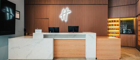 A reception desk with a wooden finish and a marble base, a neon sign above, and a snack display to the right.