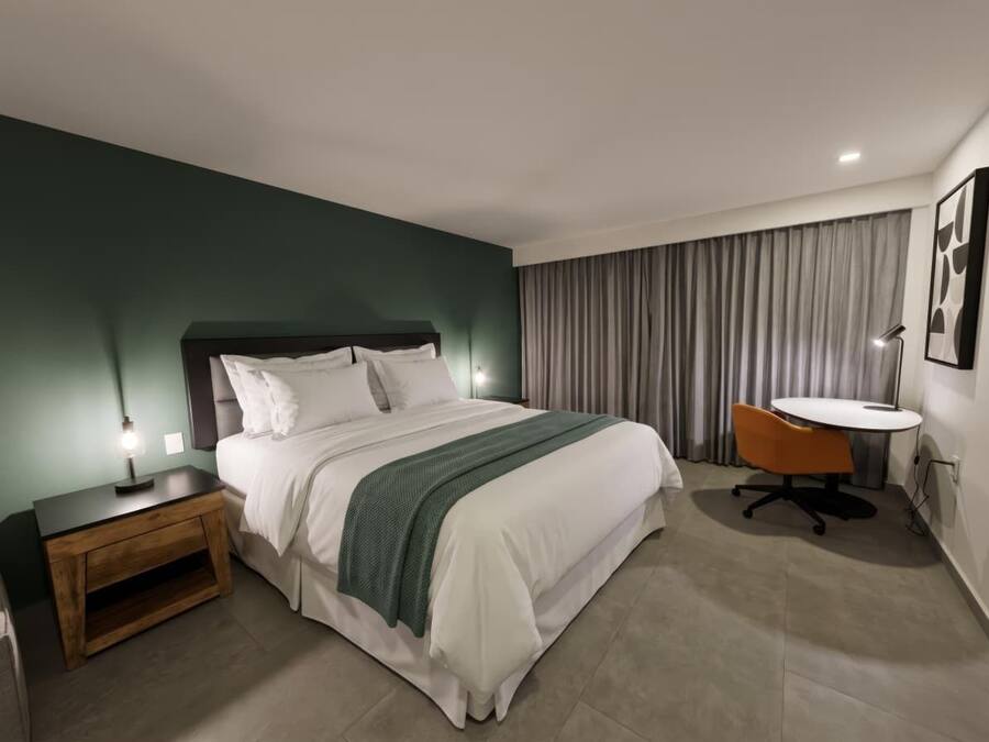 A hotel room with a bed, bedside table, lamp, chair, and a small table with a lamp.