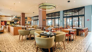 A spacious hotel dining area with multiple tables and chairs, a central column, and large windows providing views.