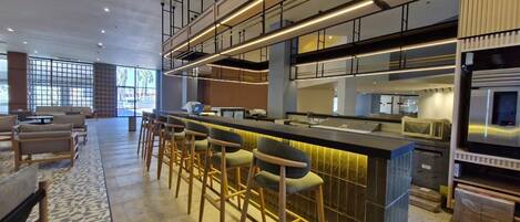 A modern bar area with a long counter and multiple stools, a kitchen with stainless steel appliances, and a seating area with sofas and a patterned rug.