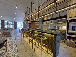 Un bar moderno con una barra larga y varios taburetes, una cocina con electrodomésticos de acero inoxidable, y un área de estar con sofás y un tapete estampado.