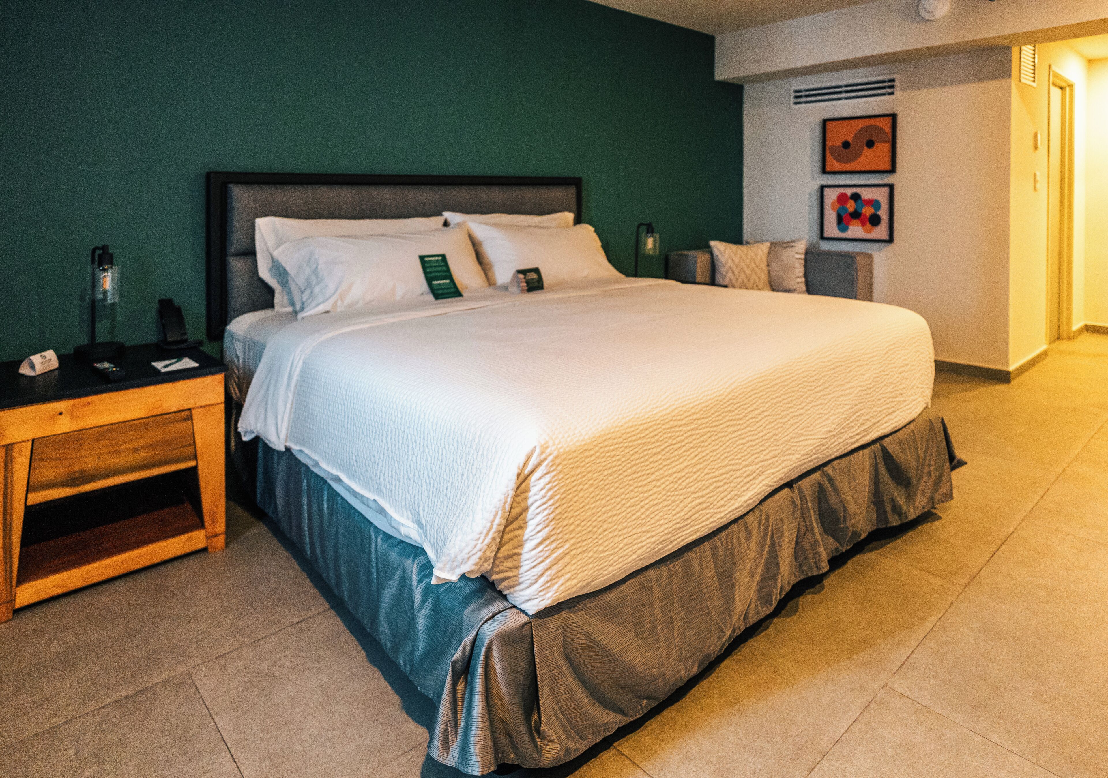 A hotel room with a bed, a nightstand, a lamp, and a wall with artwork.