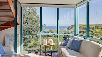 Habitación con una ventana grande que ofrece vistas al océano y colinas. Hay un sofá y una mesita con velas.