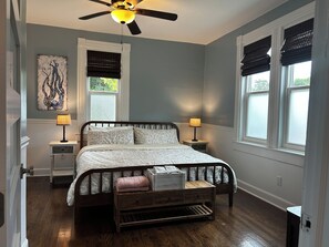 Une chambre à coucher comprenant un lit, deux tables de chevet, un ventilateur de plafond, une fenêtre avec des stores et une œuvre d’art accrochée au mur.