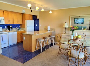 A kitchen with a central island, bar stools, a dining table with chairs, and a living area with a sofa and a painting. - Exquisite Direct Sunset & Oceanview 4 Miles! Pool (Lahaina)