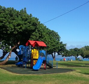A playground with a blue slide and a red roof, surrounded by trees and a body of water in the background. - Exquisite Direct Sunset & Oceanview 4 Miles! Pool (Lahaina)