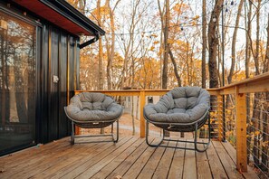 Eine Holzterrasse mit zwei grau gepolsterten Stühlen, einer Glastür und Blick auf Herbstbäume.