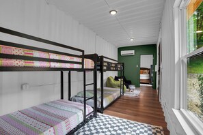A bunk bed room with a patterned bedspread, a window with a view of greenery, and a hallway leading to another room. - The Lodge at Rivers Edge Retreat | Hot tub + Pickleball (Malvern)