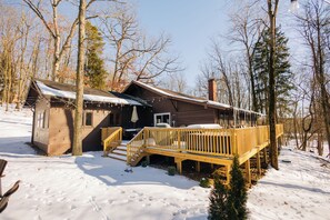 A cabin with a wooden deck and a covered porch, surrounded by snow and trees. - The Lodge at Rivers Edge Retreat | Hot tub + Pickleball (Malvern)