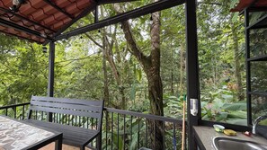 A covered outdoor seating area with a bench and table, surrounded by lush greenery and trees.