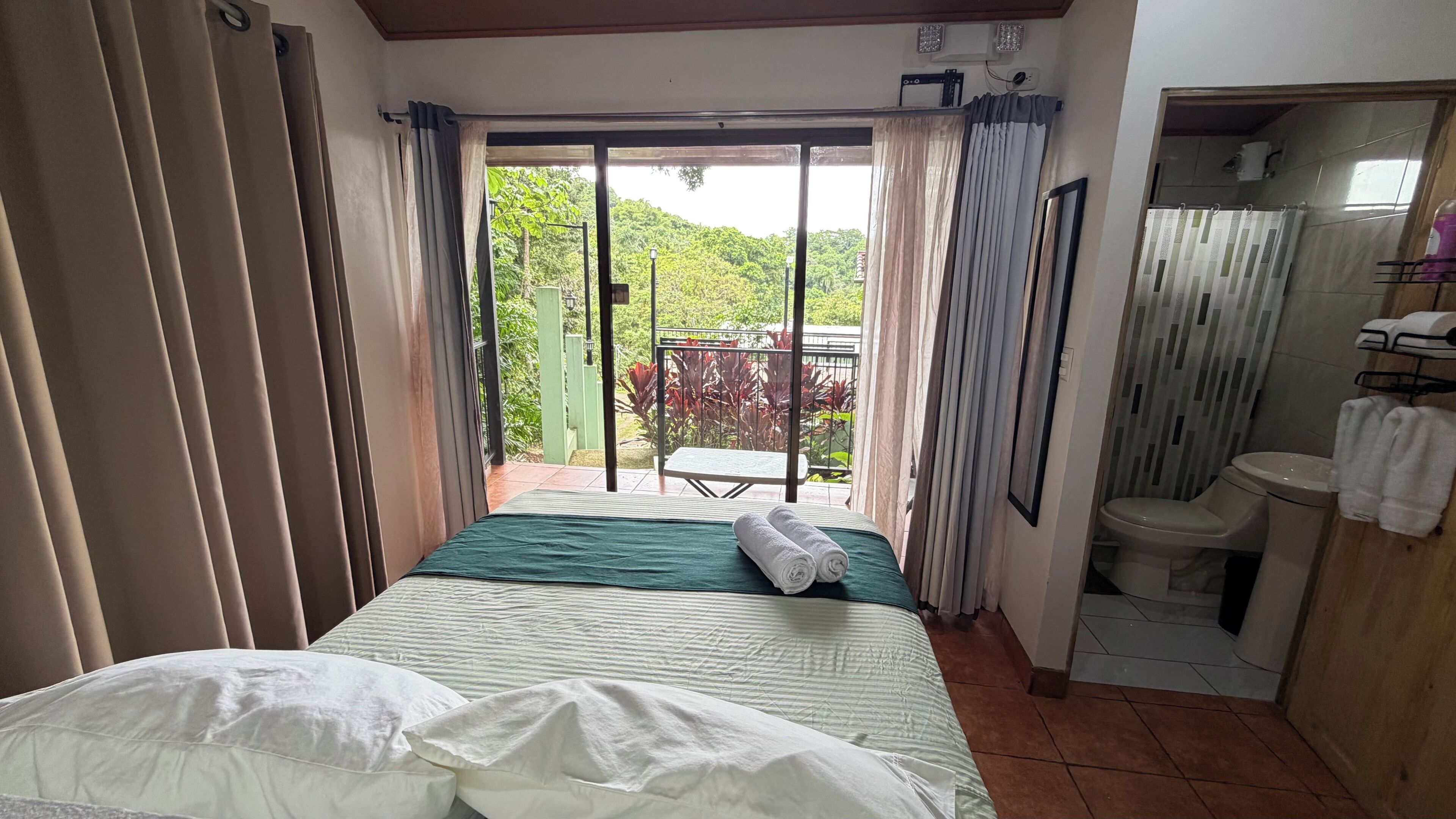 A room with a bed, a small table, a bathroom, and a view of greenery outside.