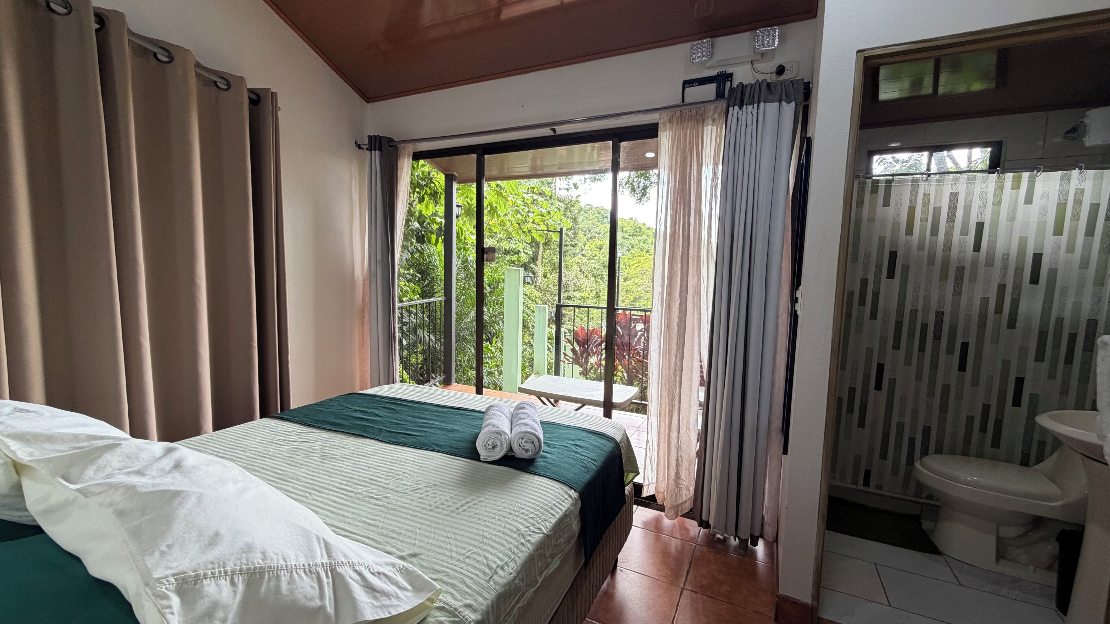 A hotel room with a bed, a view of greenery, a bathroom with a toilet and sink, and curtains.