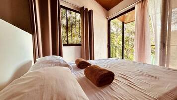 A hotel room with a bed, two rolled towels, and a view of greenery through the windows.