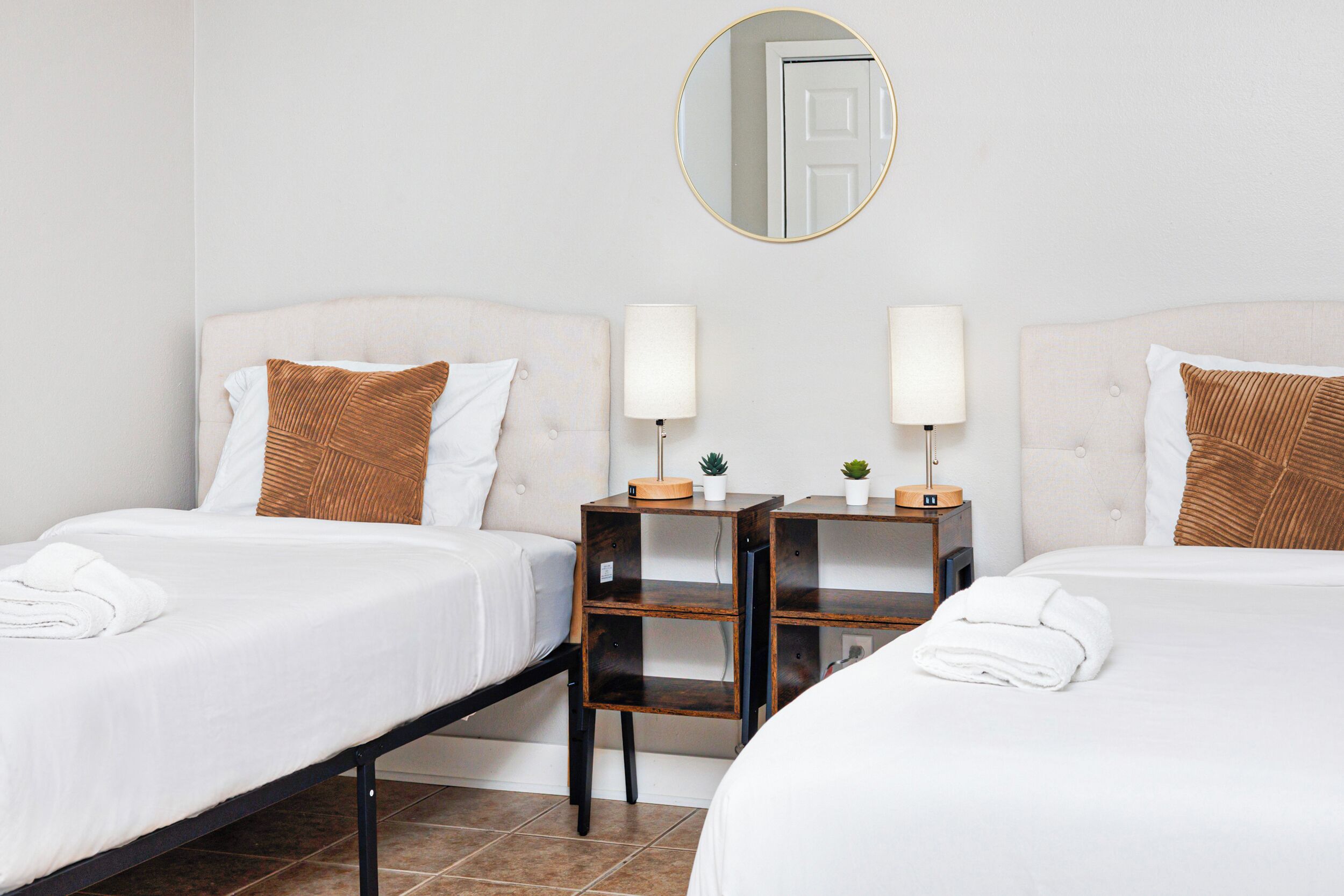 Een hotelkamer met twee bedden, elk voorzien van een witte dekbedovertrek, een bruin kussen en een nachtkastje met een lamp en een kleine plant.