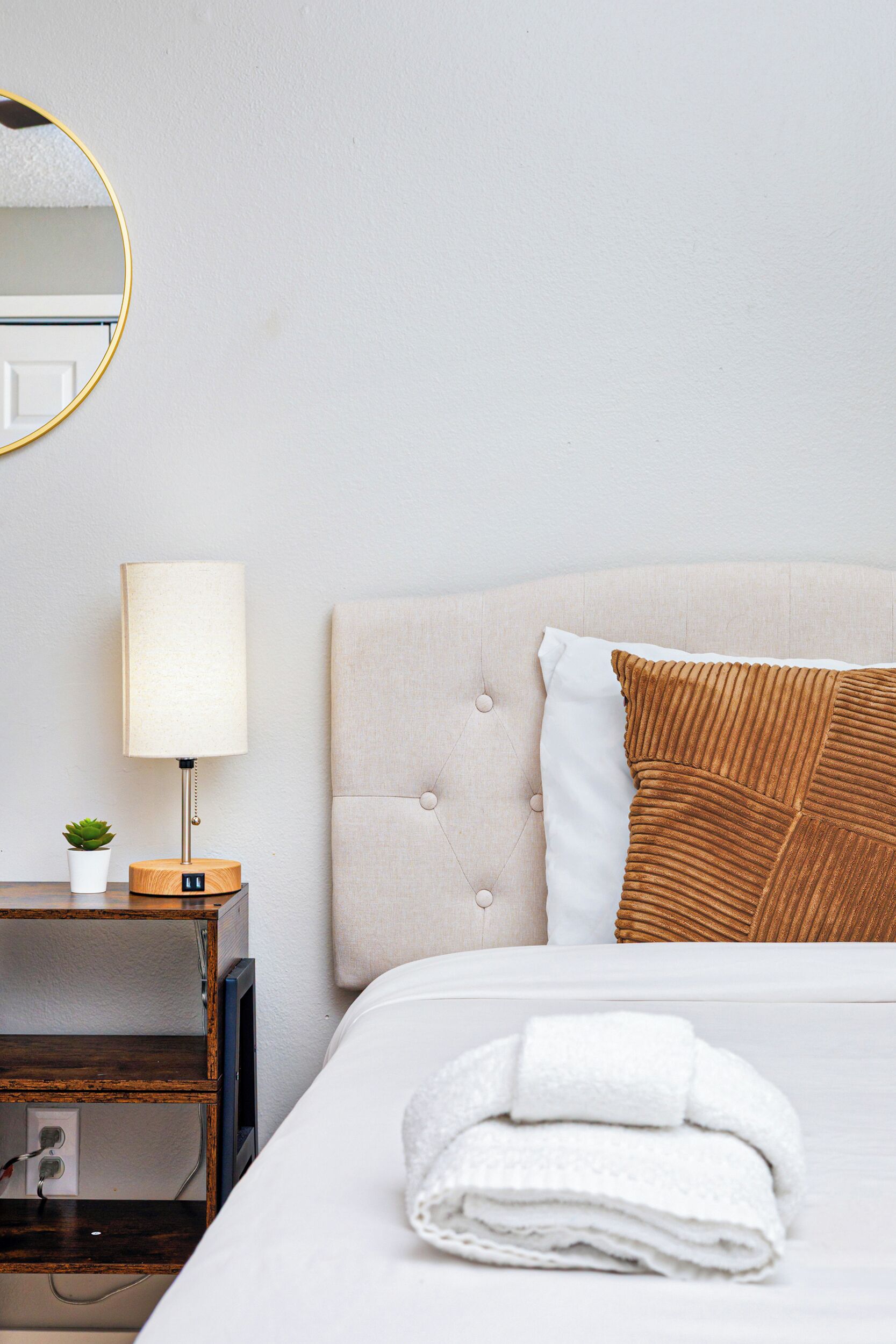 A hotel room with a bed, a bedside table with a lamp, a small plant, and a folded towel.