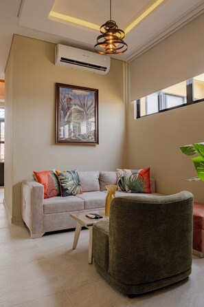 Ein modernes Wohnzimmer mit Sofa, Sesseln und Couchtisch. An der Wand hängt ein gerahmtes Bild, eine Klimaanlage ist vorhanden, und ein großes Fenster lässt viel natürliches Licht herein.
