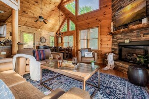 A cozy cabin interior with a fireplace, wooden walls, a dining area, and a large window.