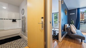 Quarto de hotel com banheira, porta de madeira, sofá cinzento e vista para o exterior.