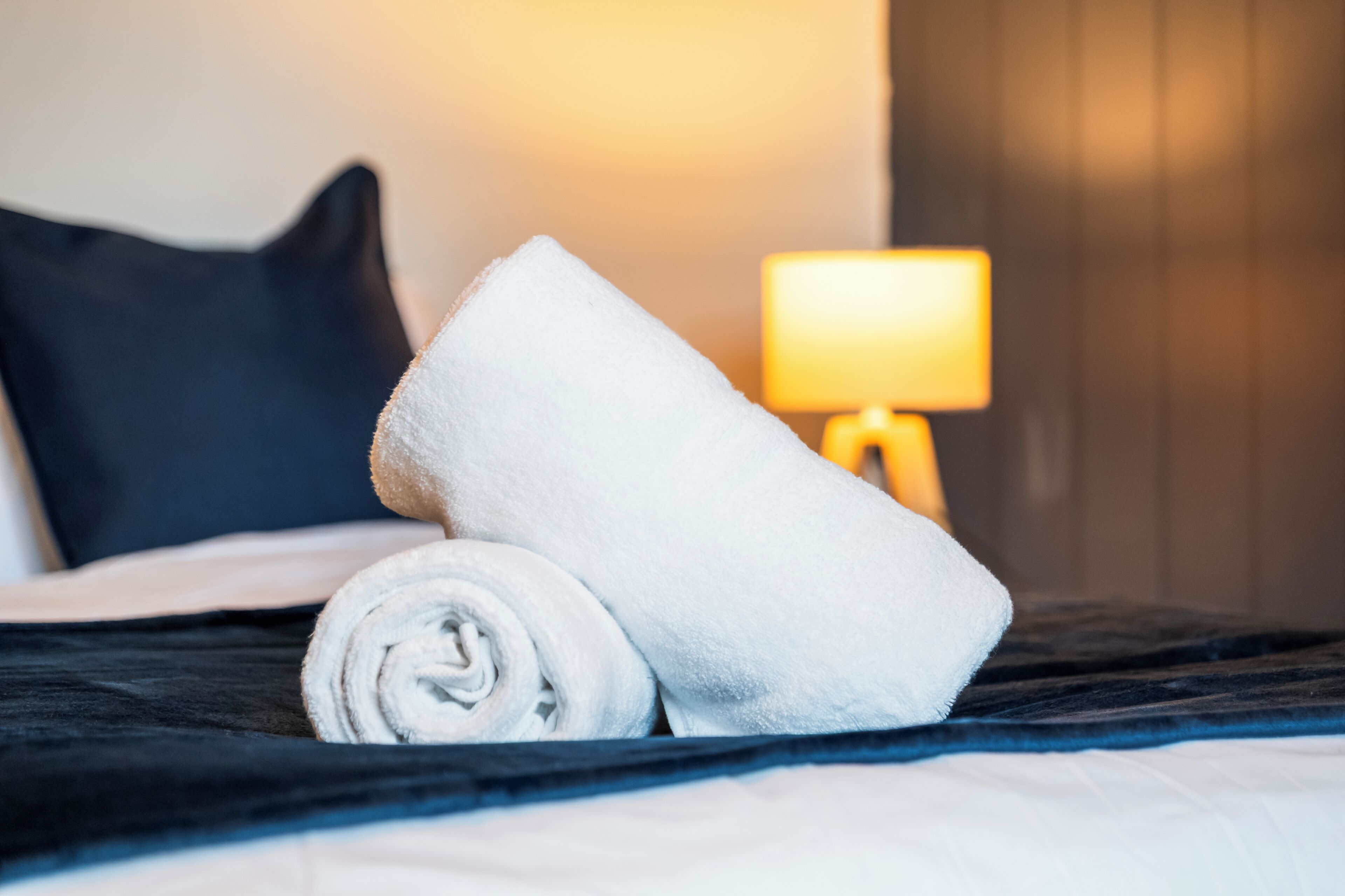 Een net opgemaakt bed met een opgerolde witte handdoek, een zwart kussen en een bedlamp.