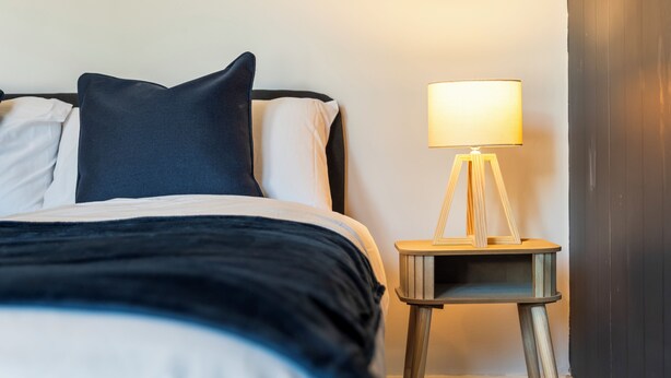 Uma cama com cabeceira escura, travesseiros brancos e escuros, e uma luminária de cabeceira sobre uma pequena mesa de cabeceira de madeira.