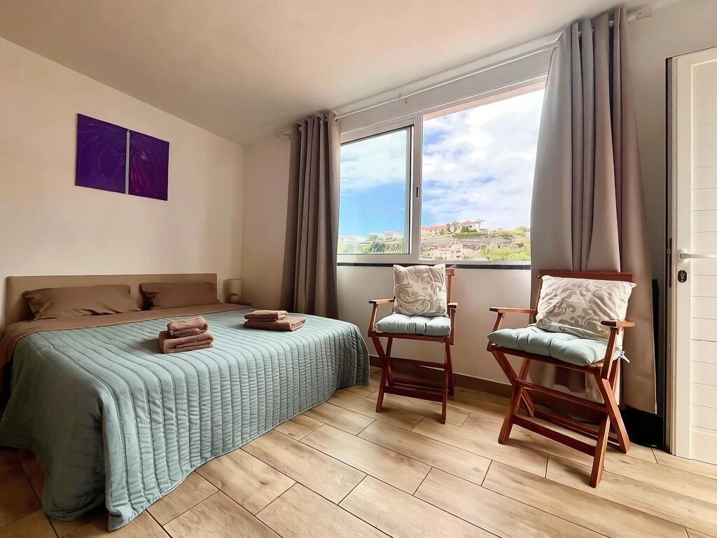 Ein Schlafzimmer mit einem Bett, zwei Stühlen, einem Fenster mit Vorhängen und Blick nach draußen.