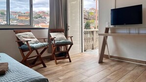 A room with a bed, two chairs, a television, and a balcony with a view of buildings. - Recharge in this peaceful and stylish place. (Funchal)