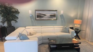 Uma sala de estar moderna com um sofá seccional, uma mesa de centro de vidro, uma planta em vaso e um quadro com uma paisagem.